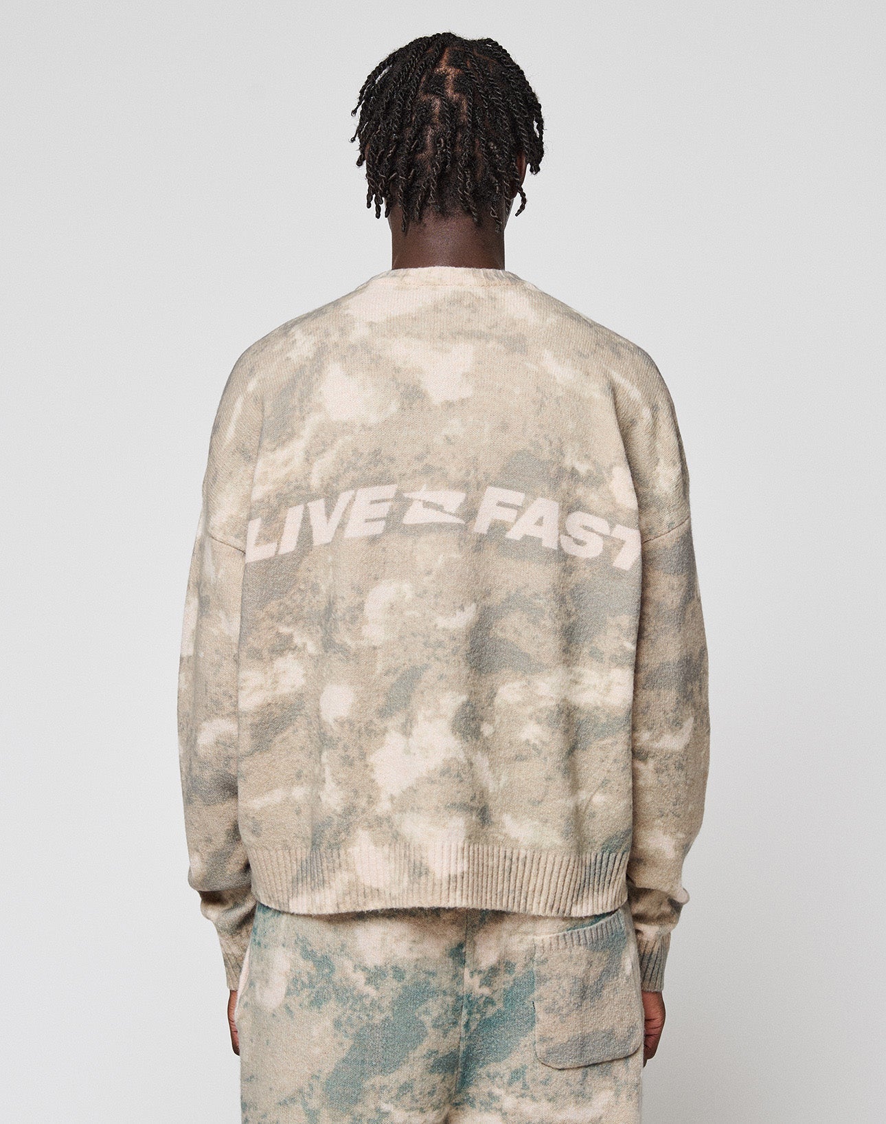 A person with short twisted hair faces away from the camera, wearing the LIVE FAST DIE YOUNG Live Fast Boucle Sweater—an oversized beige and gray tie-dye top with "LIVE FAST" boldly across the back—and matching polyester pants.