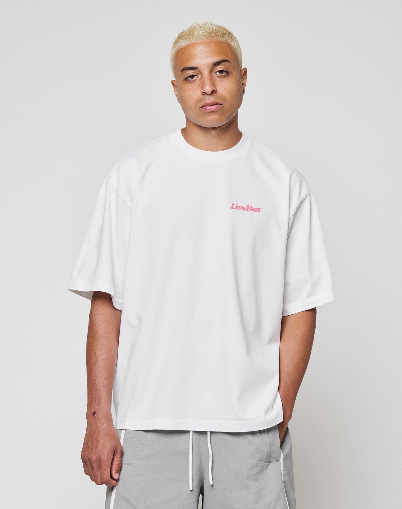 A person with short bleached blond hair wears the LIVE FAST DIE YOUNG Basic 280 Tee, a loose white 100% Baumwolle T-shirt with small pink “LiveFast” text on the chest, plus light gray drawstring shorts, standing against a plain light background.