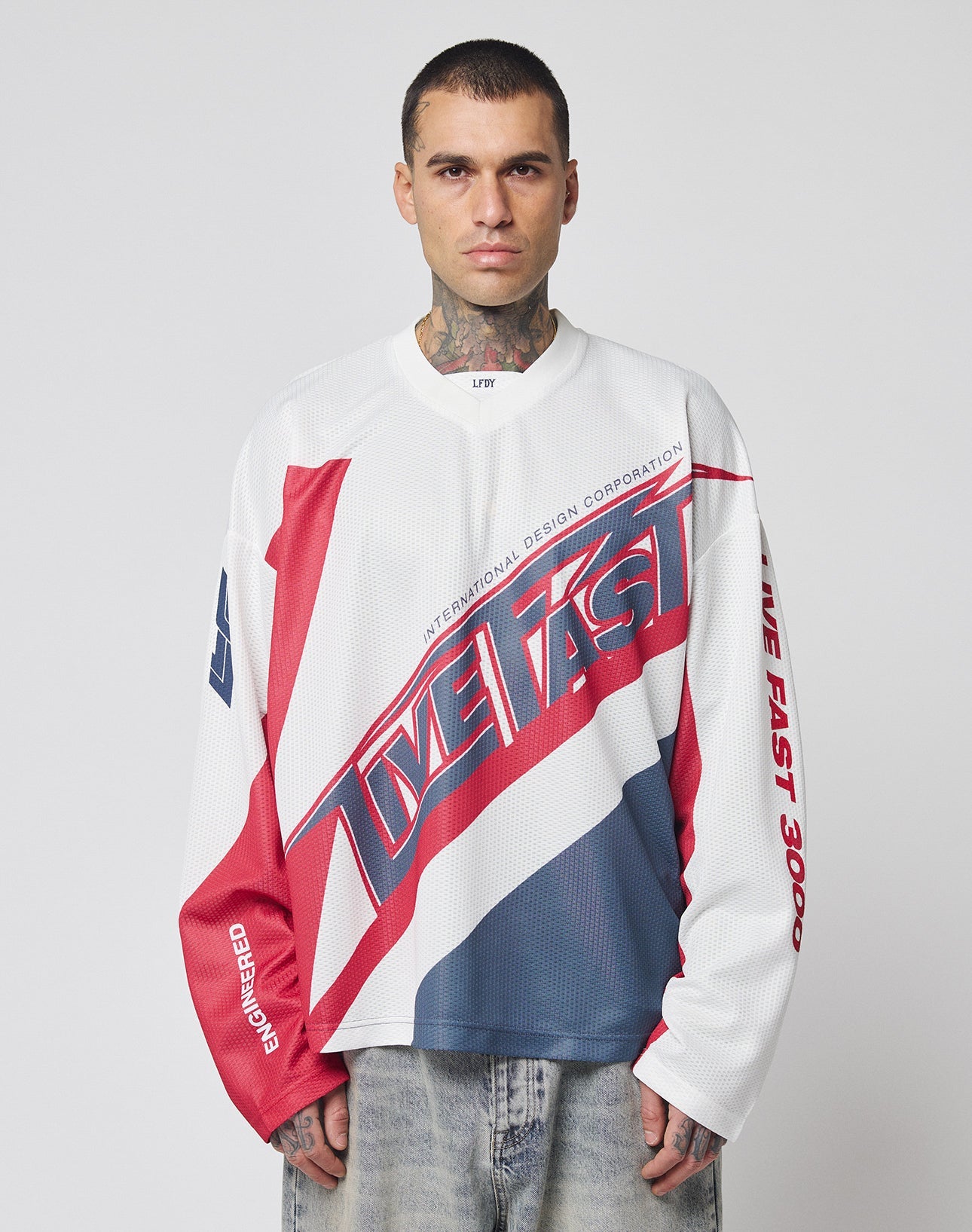 A man with short hair and neck tattoos wears the LFDY Racetrack Jersey by LIVE FAST DIE YOUNG—an oversized, long-sleeve 100% polyester shirt with bold red, white, and blue multi-logo prints and LIVE FAST text. He stands against a light gray background.