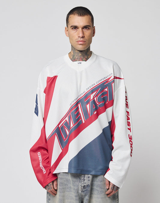 A man with short hair and neck tattoos wears the LFDY Racetrack Jersey by LIVE FAST DIE YOUNG—an oversized, long-sleeve 100% polyester shirt with bold red, white, and blue multi-logo prints and LIVE FAST text. He stands against a light gray background.