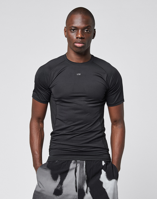 A man stands facing forward, wearing the LIVE FAST DIE YOUNG LFDY Performance Slim Tee with a reflective "LØW" chest logo and black-and-white patterned shorts, against a plain light gray background.