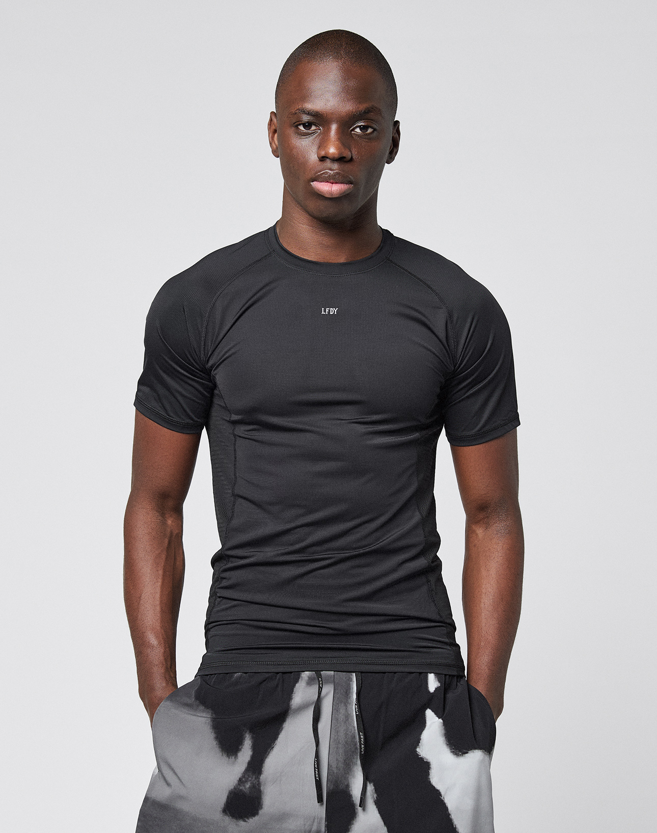 A man stands facing forward, wearing the LIVE FAST DIE YOUNG LFDY Performance Slim Tee with a reflective "LØW" chest logo and black-and-white patterned shorts, against a plain light gray background.