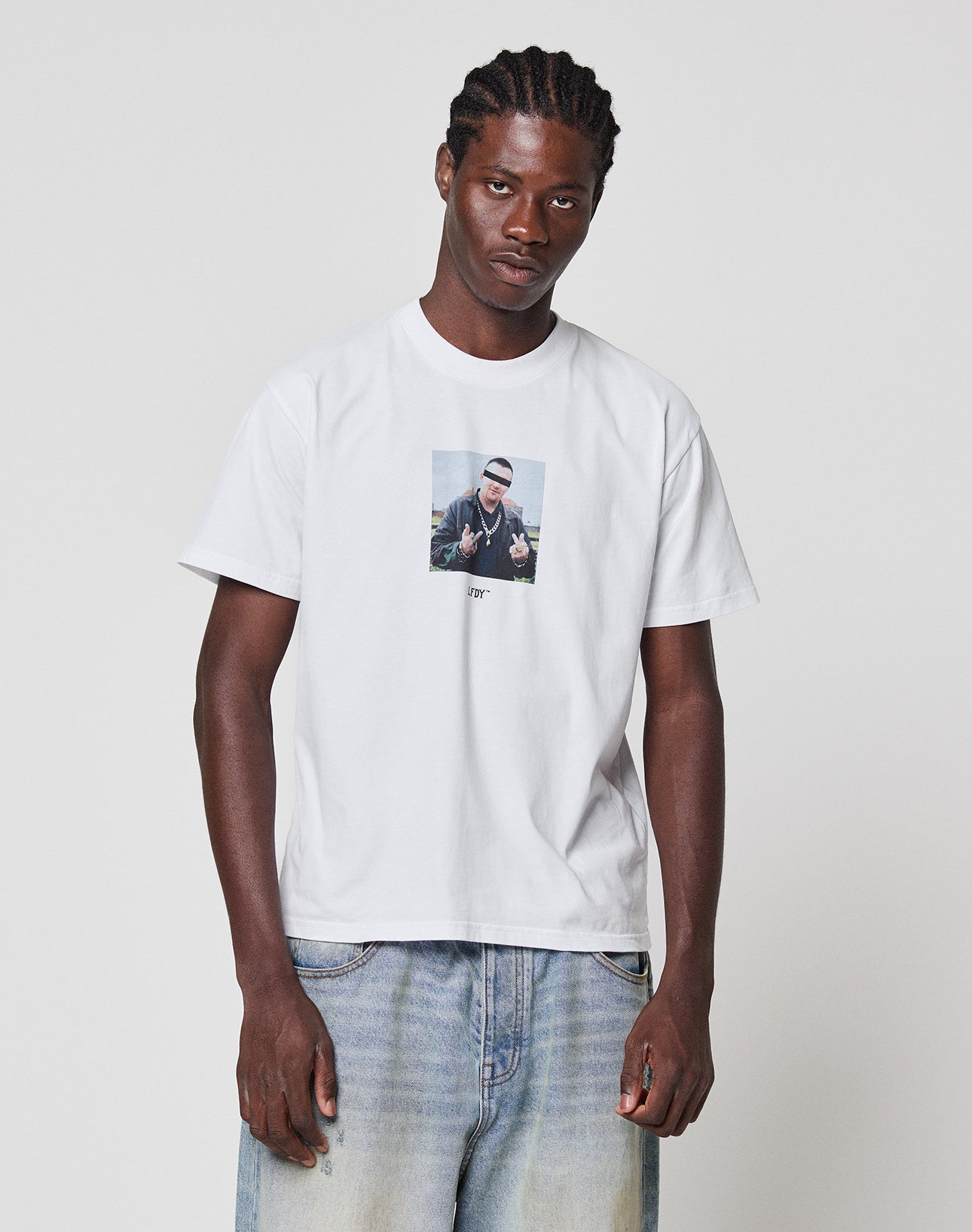 A man with braided hair wears the LIVE FAST DIE YOUNG LFDY Gambling Tee, a regular fit white t-shirt with a front photo print, paired with light blue jeans, standing against a plain white background.