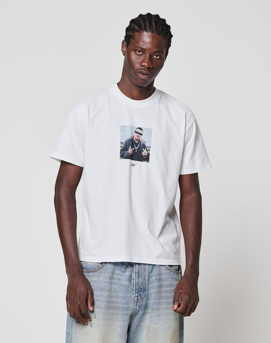 A man with braided hair wears the LIVE FAST DIE YOUNG LFDY Gambling Tee, a regular fit white t-shirt with a front photo print, paired with light blue jeans, standing against a plain white background.