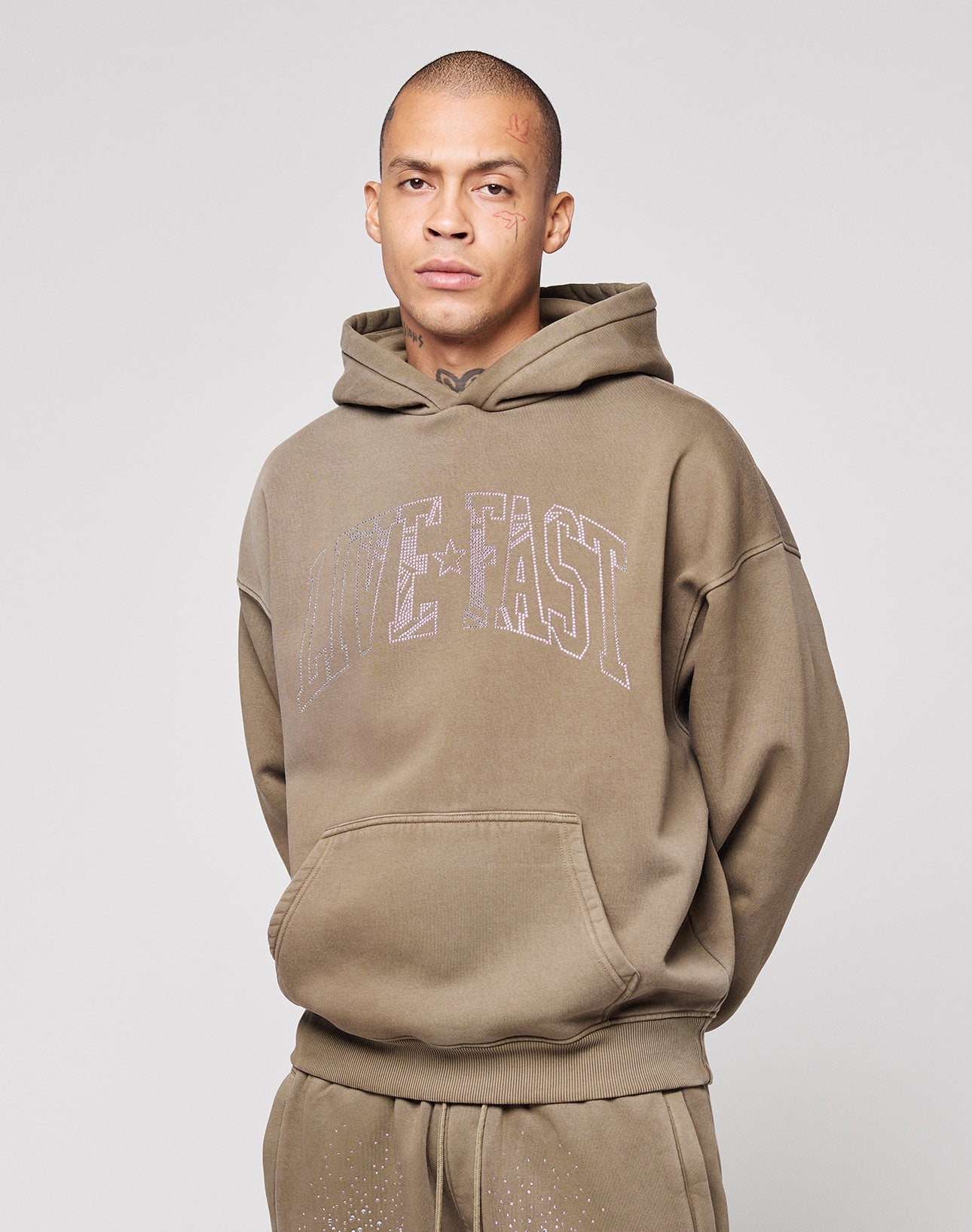 A person with short hair and face tattoos wears the LIVE FAST DIE YOUNG Live Fast Rhinestone Hooded, an oversize brown hoodie with "LIVE FAST" on the front, plus matching sweatpants. They stand against a plain light gray background, expression neutral.