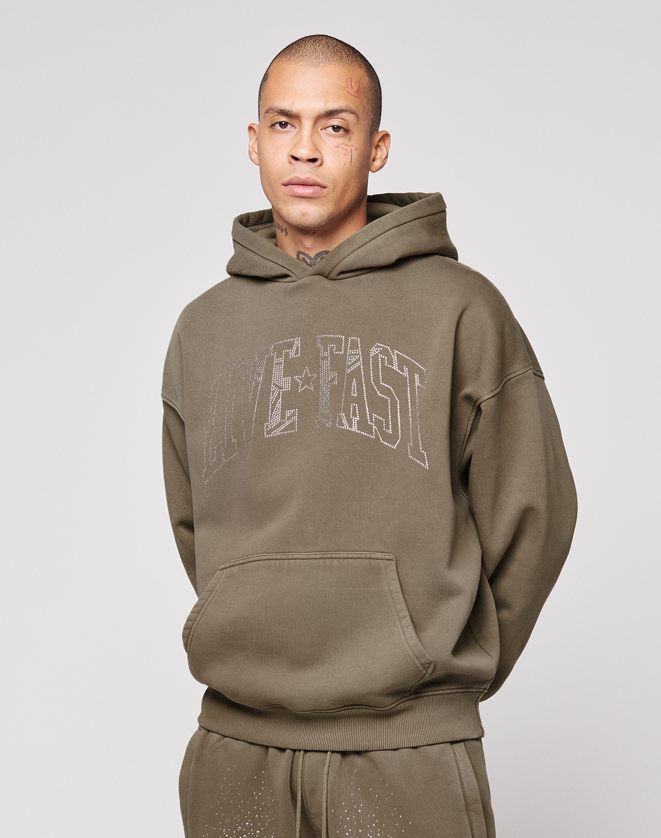 A person with short hair and face tattoos wears the LIVE FAST DIE YOUNG "Live Fast Rhinestone Hooded" in olive green, featuring a rhinestone logo and LIVE FAST print on the front, with a vintage wash and an oversized fit, against a light gray background.