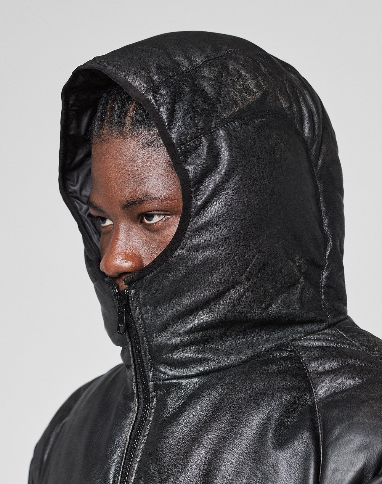 A person with braided hair zips the Live Fast Leather Puffer Jacket by LIVE FAST DIE YOUNG up to their nose, revealing only their eyes and forehead, while the embossed logo stands out against a plain light background.