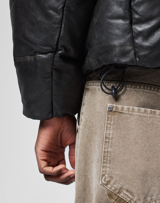 A close-up shows a person in the LIVE FAST DIE YOUNG Live Fast Leather Puffer Jacket and beige pants, their hand near the pocket with a black cord looped through a belt loop.