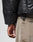 A close-up shows a person in the LIVE FAST DIE YOUNG Live Fast Leather Puffer Jacket and beige pants, their hand near the pocket with a black cord looped through a belt loop.