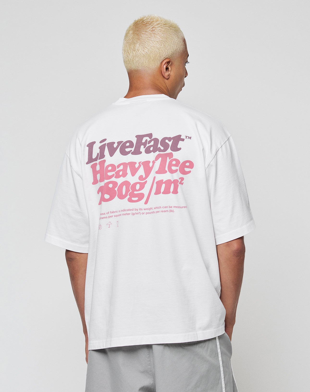 A person with short blond hair wears the LIVE FAST DIE YOUNG Basic 280 Tee—an oversized 100% Baumwolle white shirt with bold pink Live Fast Heavy Tee 180g/m² front and back print—paired with light gray shorts, facing away from the camera.