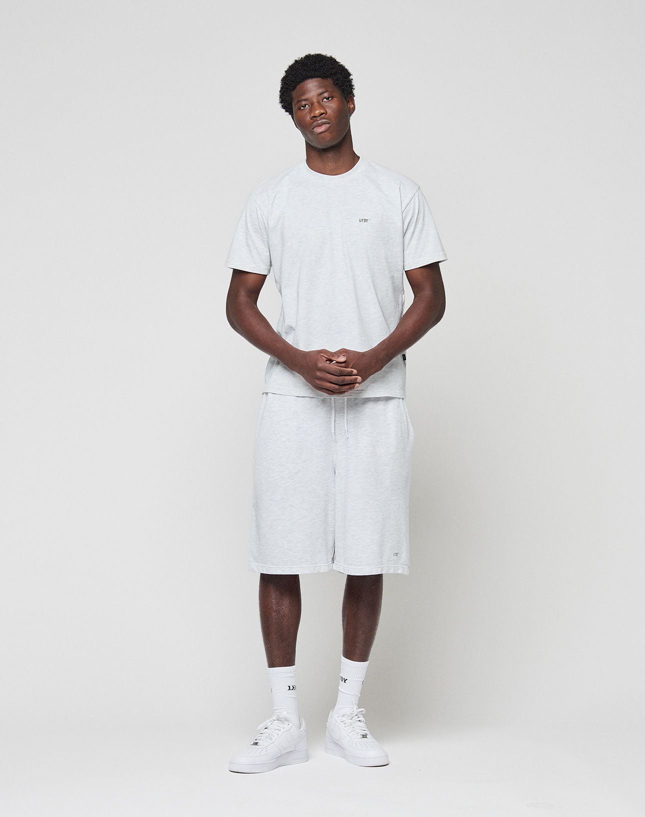 A man stands against a white background wearing the LIVE FAST DIE YOUNG Basic Chest Tee in light gray Baumwolle with matching Regular Fit shorts. He pairs the outfit with white socks and sneakers, hands clasped, gazing slightly to the side.