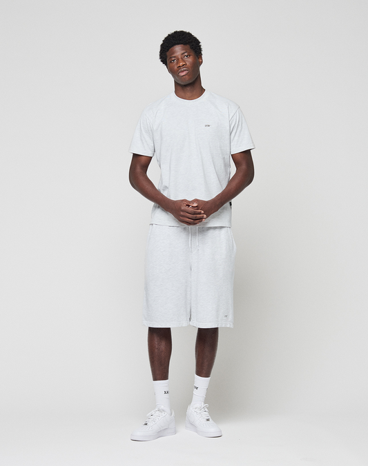 A man stands against a white background wearing the LIVE FAST DIE YOUNG Basic Chest Tee in light gray Baumwolle with matching Regular Fit shorts. He pairs the outfit with white socks and sneakers, hands clasped, gazing slightly to the side.