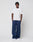 A person with short, twisted hair stands against a plain background, hands in pockets, wearing the LIVE FAST DIE YOUNG Live Fast Paris Tee (regular fit, 100% Baumwolle), loose navy pants, and white sneakers.