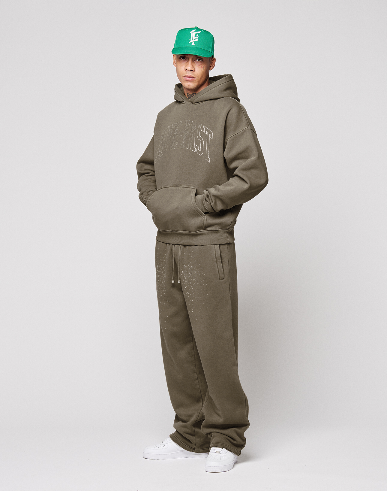 A man wears LIVE FAST DIE YOUNG's Live Fast Rhinestone Jogger in olive green, styled with a matching hoodie, bright green cap, and white sneakers. He stands against a plain background with hands in his pockets, looking at the camera.
