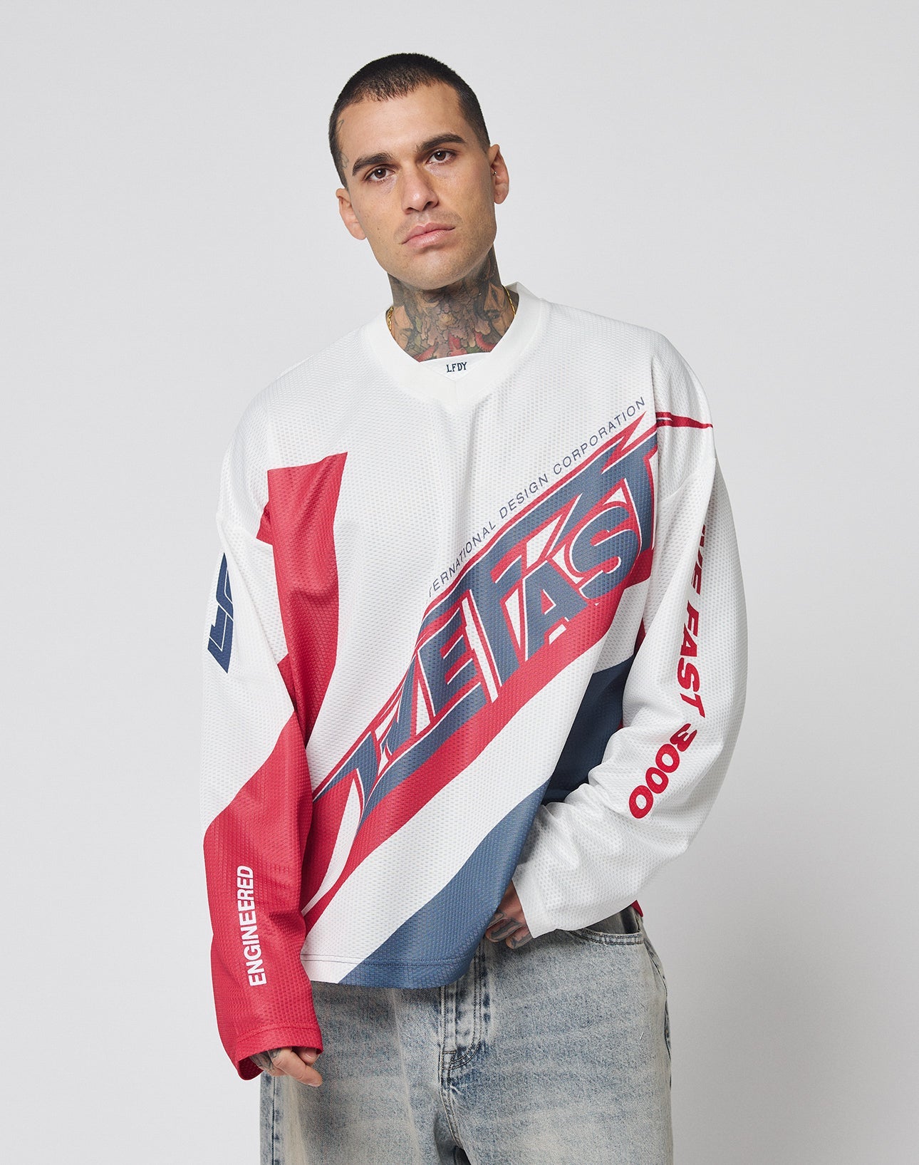 A model wears the LFDY Racetrack Jersey by LIVE FAST DIE YOUNG—a white, red, and blue long-sleeve shirt with bold multi-logo prints and “THE FAST”—posing against a plain background with one hand in their pocket.