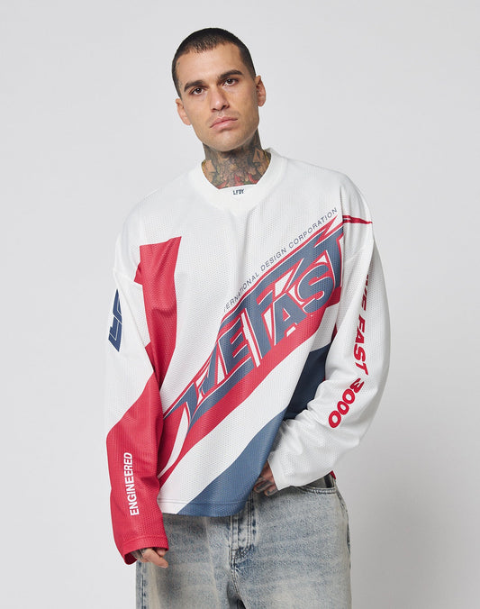 A model wears the LFDY Racetrack Jersey by LIVE FAST DIE YOUNG—a white, red, and blue long-sleeve shirt with bold multi-logo prints and “THE FAST”—posing against a plain background with one hand in their pocket.