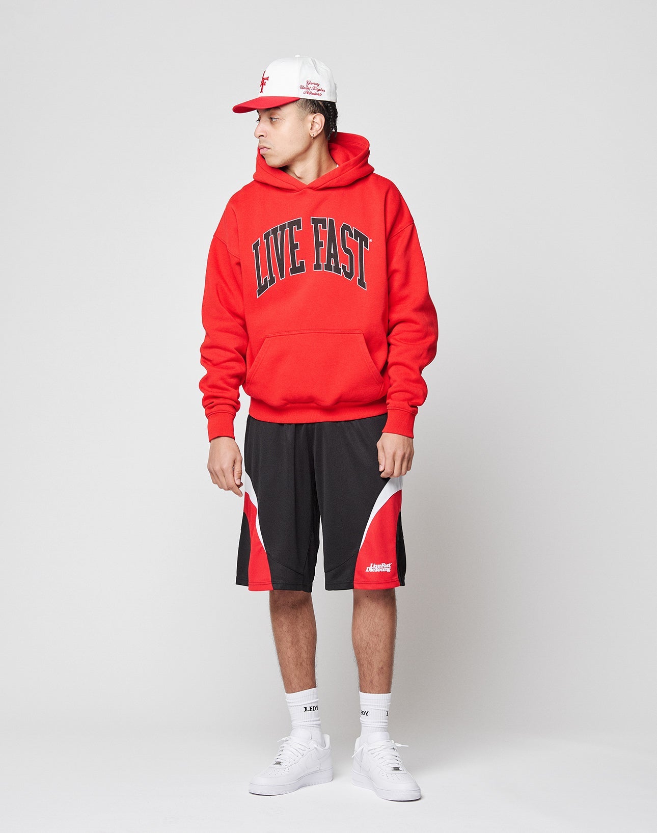A person in a red LIVE FAST DIE YOUNG Live Fast College Hooded with backprint, black and red athletic shorts, white sneakers, and a white/red cap stands against a plain white background looking to the side.