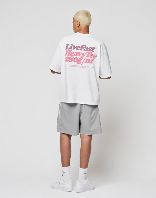 A person with short blond hair faces away, wearing the LIVE FAST DIE YOUNG Basic 280 Tee in oversize fit with bold pink backprint, paired with light gray shorts, white socks, and white sneakers against a plain white background.