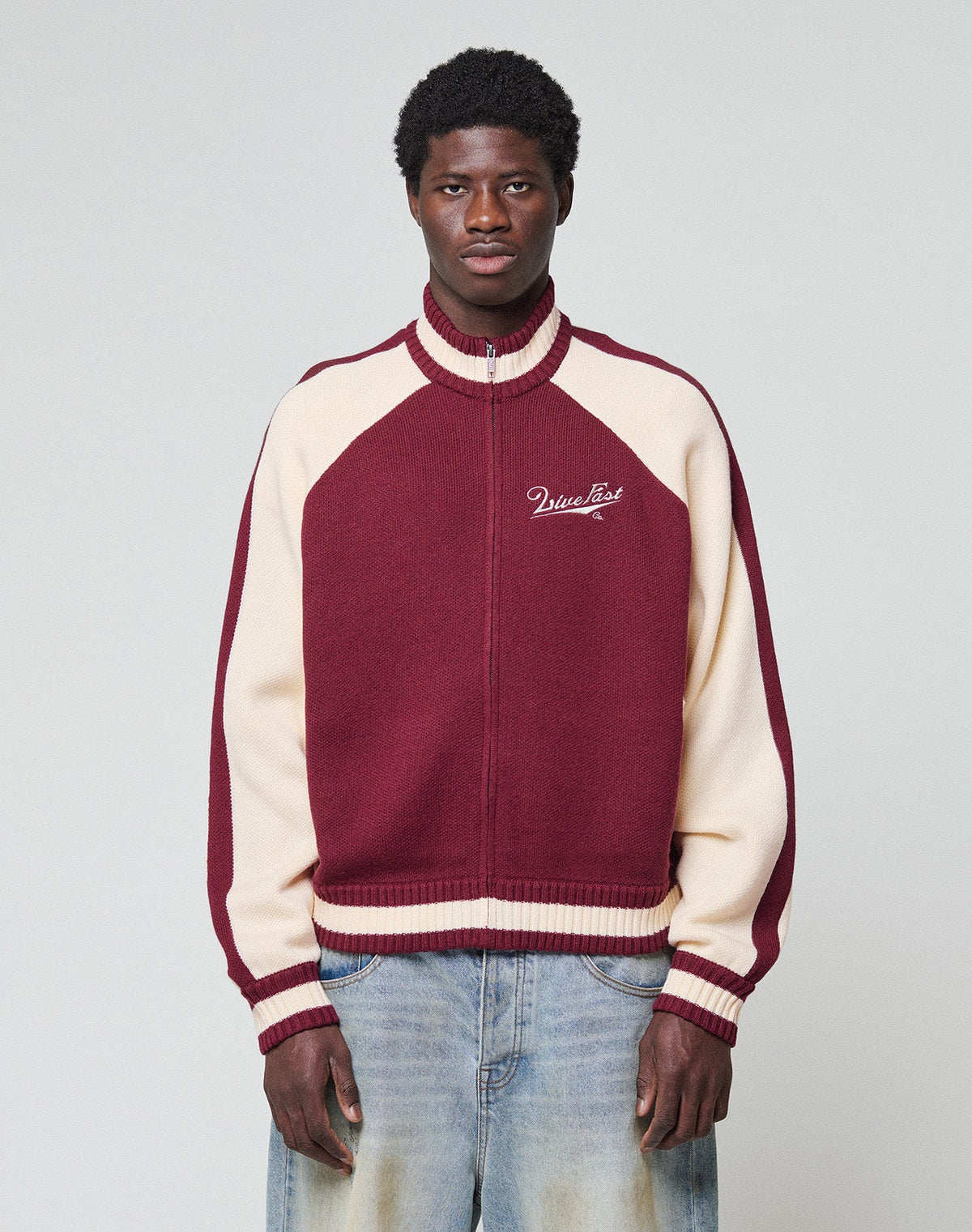 A young man with short curly hair and a neutral expression stands against a plain background, wearing the LIVE FAST DIE YOUNG Boxy Knit Zip Jacket in maroon and cream, featuring "MissList" on the chest, paired with light blue jeans.