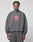 A person stands facing forward in a loose-fitting DY Sweater by LIVE FAST DIE YOUNG, made from cotton. The dark grey oversize sweatshirt displays a red and white graphic logo on the chest. The background is plain and light-colored.