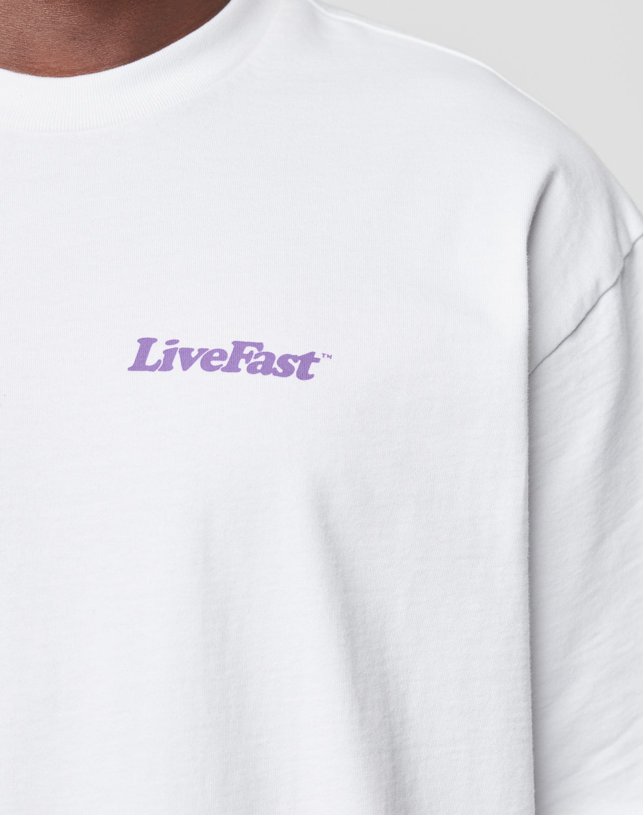 Close-up of a person wearing the LIVE FAST DIE YOUNG Basic 280 Tee, an oversize fit white T-shirt made from 100% Baumwolle, featuring “LiveFast” in light purple text on the left chest. The background is plain light gray.