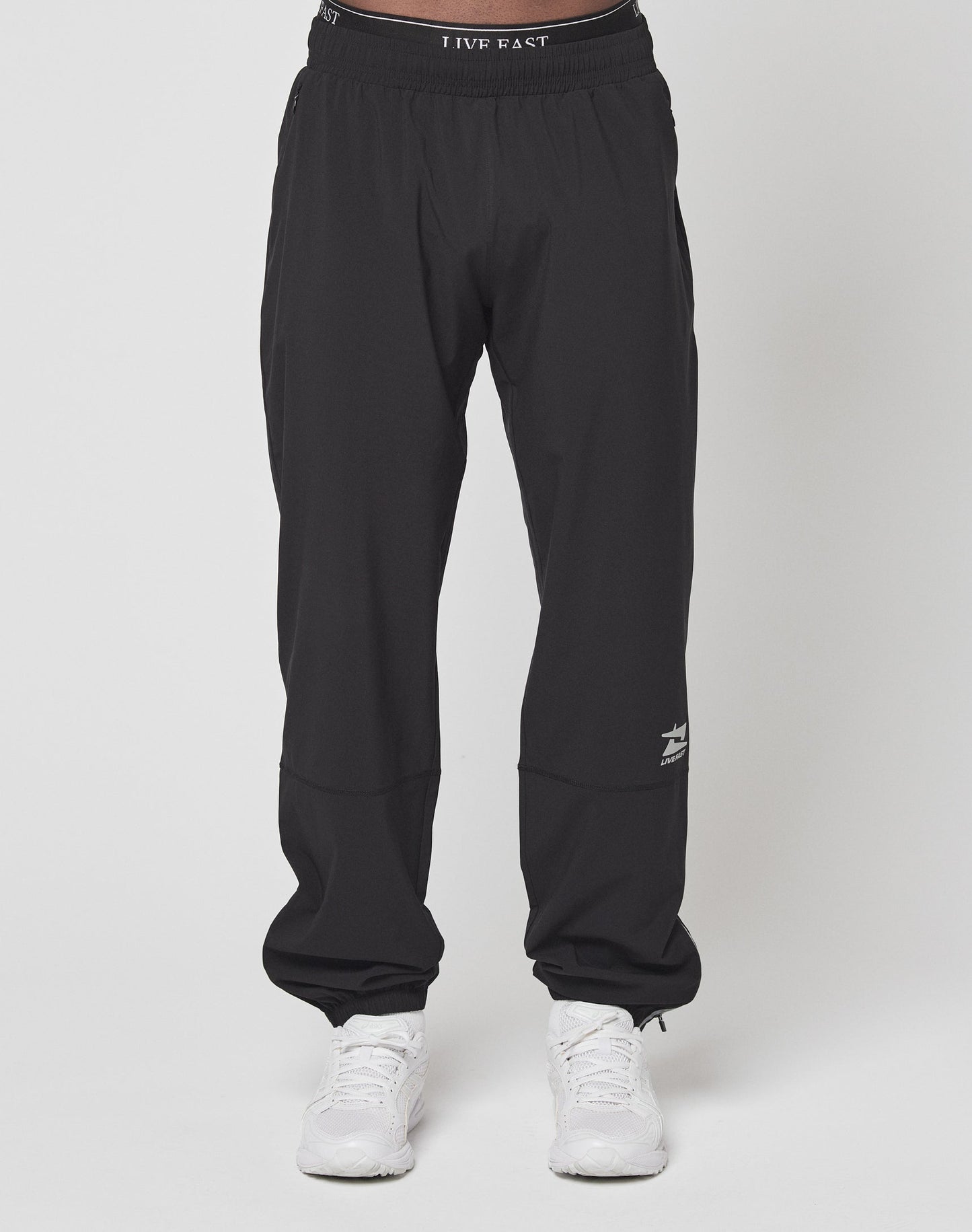 A person wears LIVE FAST DIE YOUNGs black Live Fast Performance Running Pants with a white shark logo, reflective print, and LIVE FAST text on the left leg, plus an elastic waistband, paired with white athletic shoes against a plain background.| model