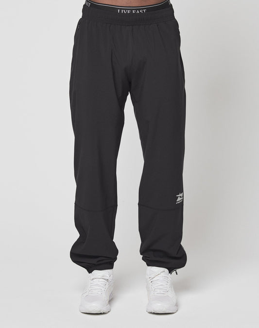 A person wears LIVE FAST DIE YOUNGs Live Fast Performance Running Pants—black joggers with a reflective print by the left knee, an elastic waistband for comfort—and white sneakers, standing against a plain white background.