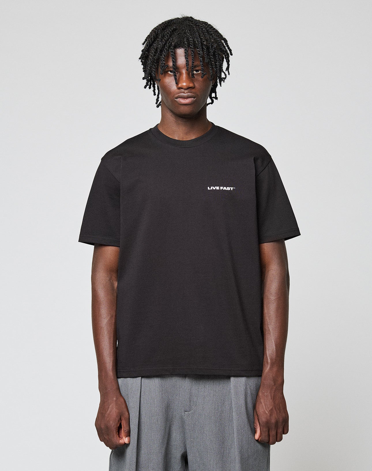 A person with short, twisted hair wears the LIVE FAST DIE YOUNG Live Fast Plain Tee—regular fit, black, 100% Baumwolle with a subtle white LIVE FAST frontprint—paired with gray pants against a plain light background.