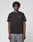 A person with short, twisted hair wears the LIVE FAST DIE YOUNG Live Fast Plain Tee—regular fit, black, 100% Baumwolle with a subtle white LIVE FAST frontprint—paired with gray pants against a plain light background.