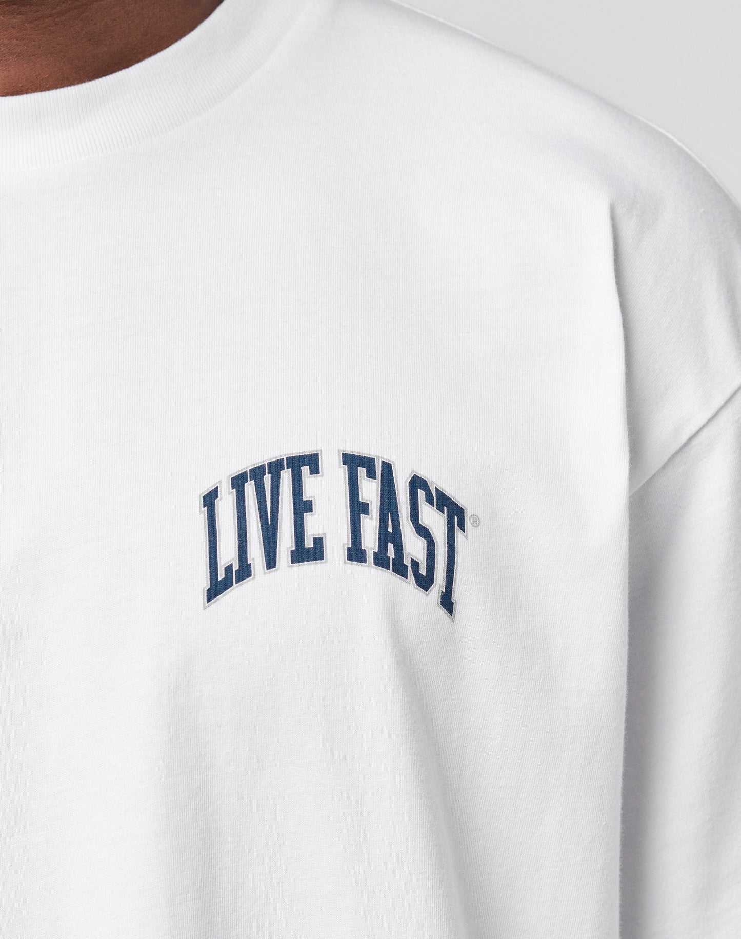 Close-up of a person wearing the Live Fast London Tee by LIVE FAST DIE YOUNG—a white, 100% cotton t-shirt with bold, curved navy LIVE FAST lettering on the chest. The Oversize Fit offers extra comfort and style.