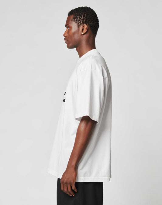 A person with short curly hair stands in profile against a plain background, wearing the oversize fit Fuck Die Young Tee by LIVE FAST DIE YOUNG, made from 100% Baumwolle, paired with black pants.