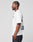 A man with braided hair wears the LIVE FAST DIE YOUNG “Live Fast London Tee,” a white oversized t-shirt with dark front and back prints, paired with dark jeans. He stands in profile against a plain white background.