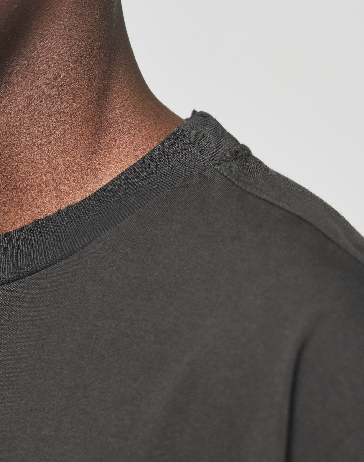 Close-up of a person wearing the LIVE FAST DIE YOUNG LFDY Sharkhunter Tee, a dark 100% Baumwolle crew neck with individuelle Distressings at the frayed neckline, showing part of their neck and shoulder against a plain background.