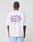 A person with braided hair is seen from behind, wearing a white Basic 280 Tee by LIVE FAST DIE YOUNG with purple Front- und Backprint text and smaller details, paired with light blue shorts.