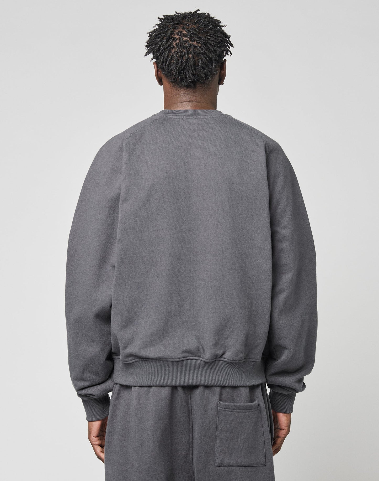 A person with short, twisted hair is seen from the back wearing a loose-fitting LIVE FAST DIE YOUNG DY Sweater and matching gray pants with a pocket, standing against a plain light background.