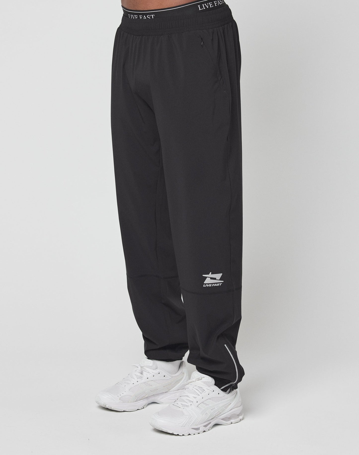 A person wears LIVE FAST DIE YOUNGs black Live Fast Performance Running Pants with a white shark logo, reflective print, and LIVE FAST text on the left leg, plus an elastic waistband, paired with white athletic shoes against a plain background.