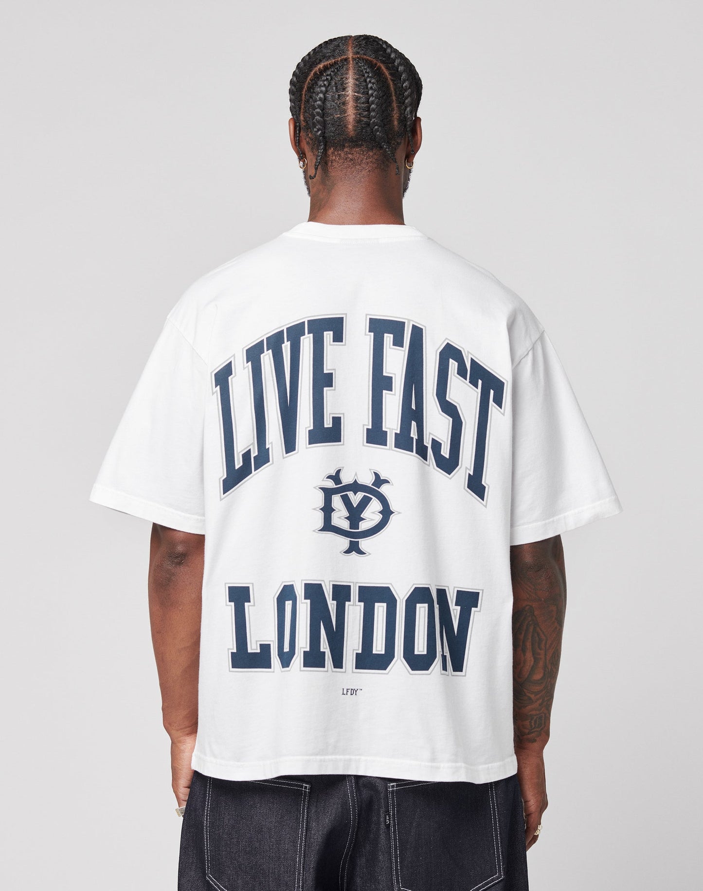 A person with braided hair faces away, wearing the LIVE FAST DIE YOUNG Live Fast London Tee, a white oversized 100% Baumwolle T-shirt featuring bold blue LIVE FAST LONDON and a circular logo on the back, paired with dark jeans. | model