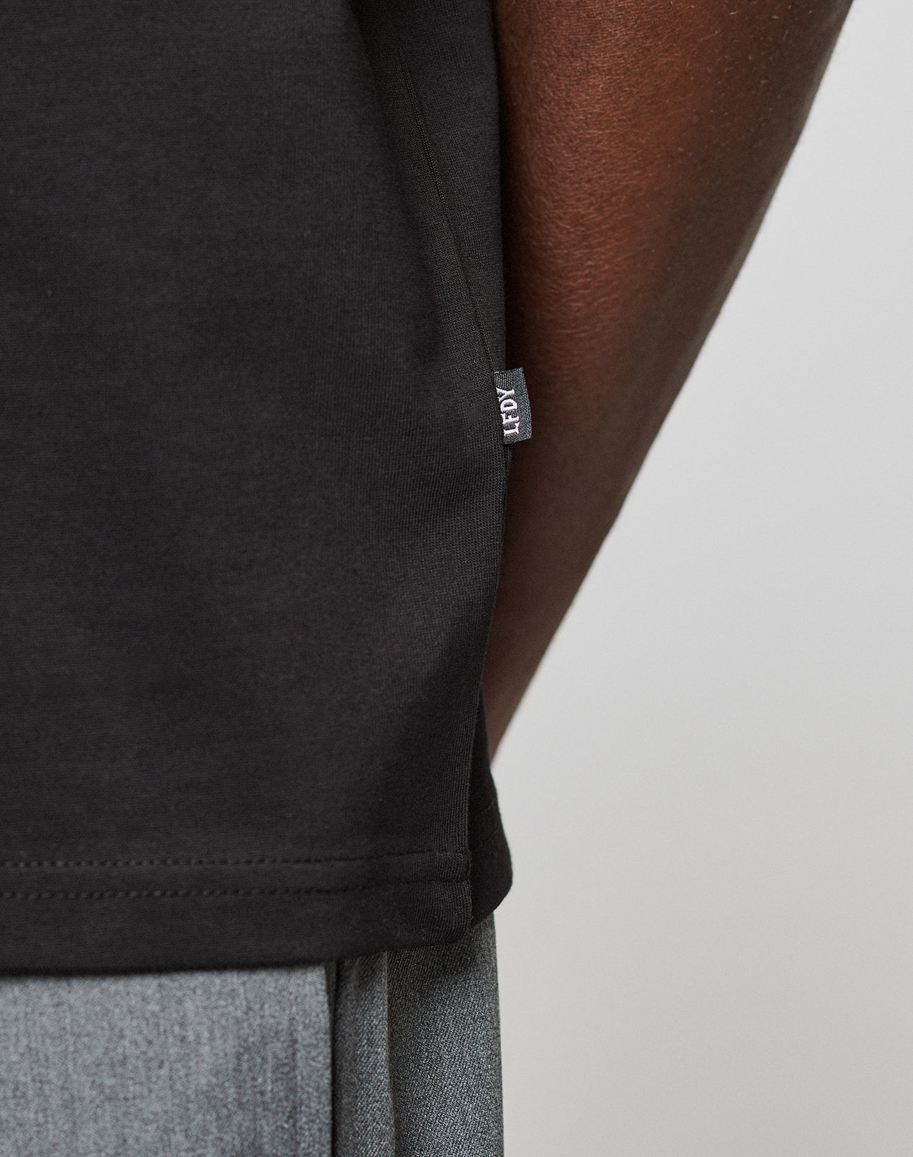 Close-up of the side of a black Live Fast Plain Tee by LIVE FAST DIE YOUNG, showing a small fabric tag. The tee is worn by a person with dark skin and gray pants visible at the bottom edge.