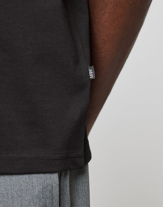 Close-up of the side of a black Live Fast Plain Tee by LIVE FAST DIE YOUNG, showing a small fabric tag. The tee is worn by a person with dark skin and gray pants visible at the bottom edge.
