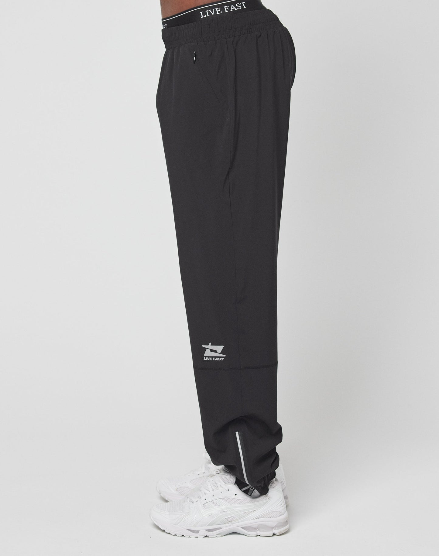 A person stands in profile against a plain white background wearing LIVE FAST DIE YOUNG’s Live Fast Performance Running Pants with an elastic waistband featuring LIVE FAST, white sneakers, and a small ankle logo.