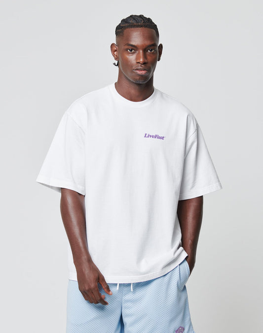 A man with short braids and a calm look wears an Oversize Fit Basic 280 Tee by LIVE FAST DIE YOUNG, made of 100% cotton with LiveFast in small purple letters, paired with light blue shorts against a plain background.