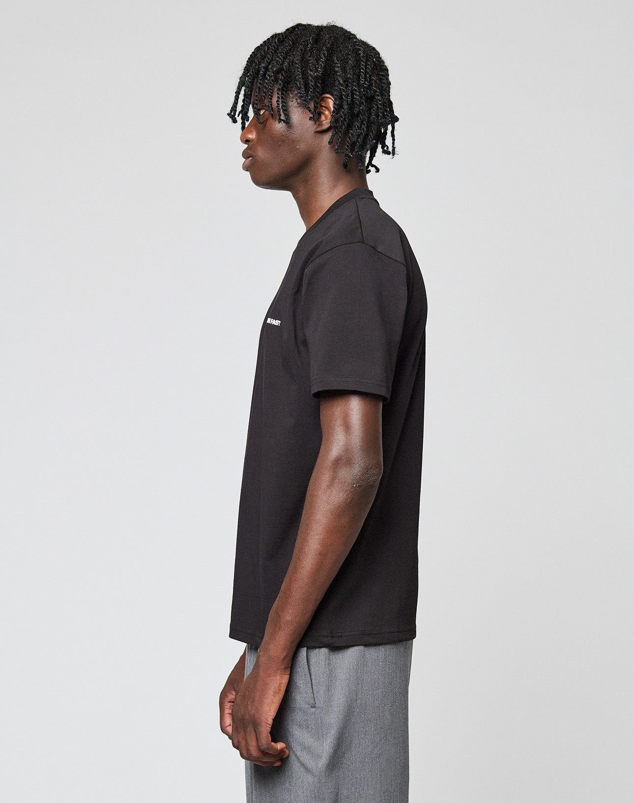 A person with short twisted hair stands in profile against a plain background, wearing a black LIVE FAST DIE YOUNG Live Fast Plain Tee with front print and gray pants.