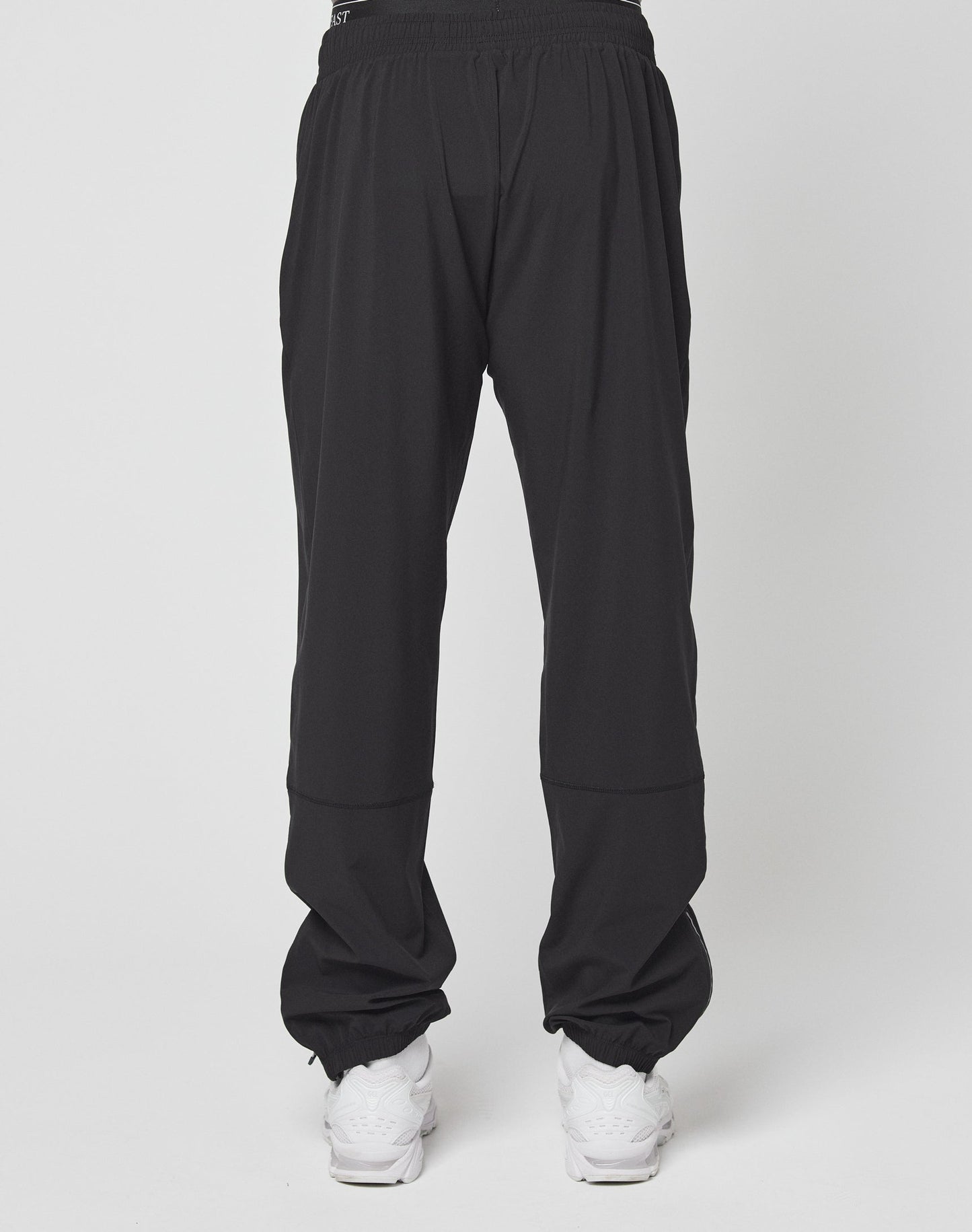 A person wears LIVE FAST DIE YOUNGs Live Fast Performance Running Pants—black joggers with a reflective print by the left knee, an elastic waistband for comfort—and white sneakers, standing against a plain white background.