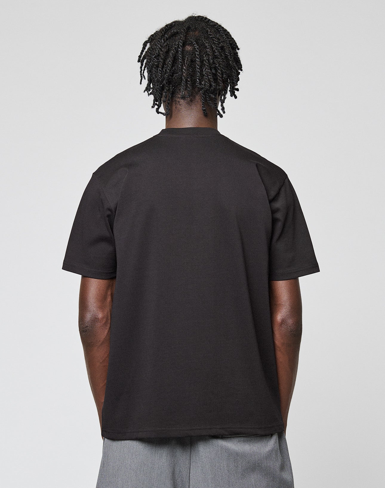 A person with short, twisted hair stands with their back to the camera, wearing the LIVE FAST DIE YOUNG Live Fast Plain Tee in black and gray shorts against a light gray background.