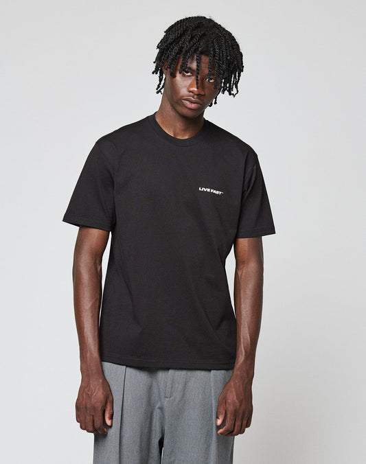 A man with short, twisted hair wears the LIVE FAST DIE YOUNG Live Fast Plain Tee in black with a front print on the left chest and loose grey pants, standing against a plain light background. Made from 100% Baumwolle.