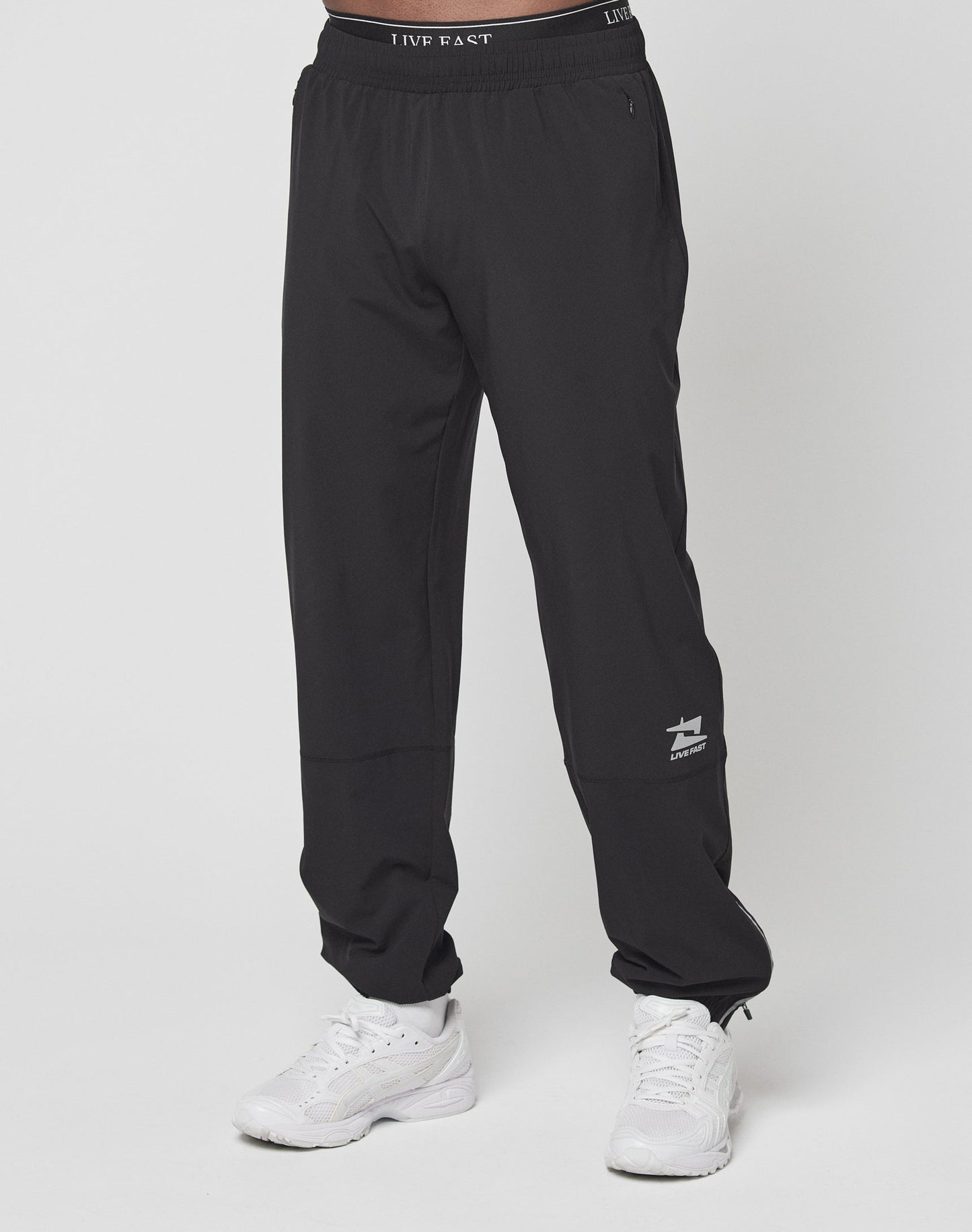 Someone is wearing the LIVE FAST DIE YOUNG Live Fast Performance Running Pants in black with reflective print, an elastic waistband, a small white logo on the left leg, and white sneakers, standing against a plain white background.