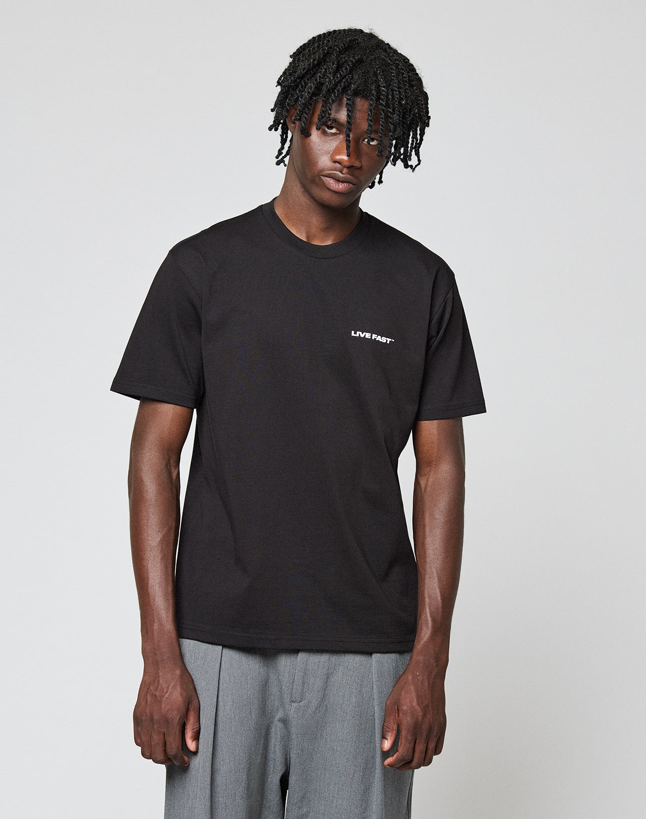 A man with short, twisted hair wears the LIVE FAST DIE YOUNG Live Fast Plain Tee in black with a front print on the left chest and loose grey pants, standing against a plain light background. Made from 100% Baumwolle.