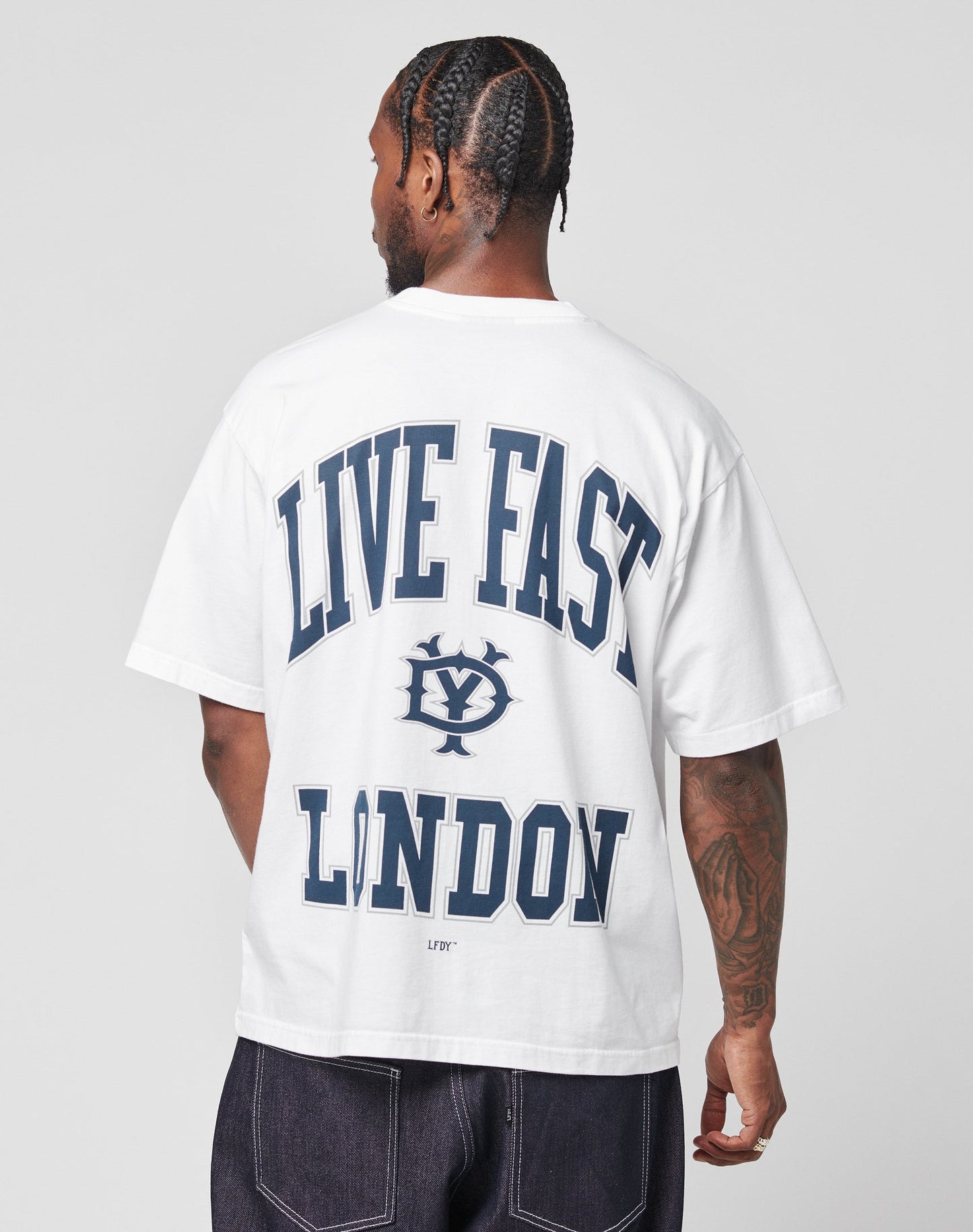 A person with braided hair faces away, wearing the LIVE FAST DIE YOUNG Live Fast London Tee—an oversize white 100% Baumwolle shirt featuring bold blue “LIVE FAST LONDON” text and a central graphic, styled with arm tattoos and dark jeans.
