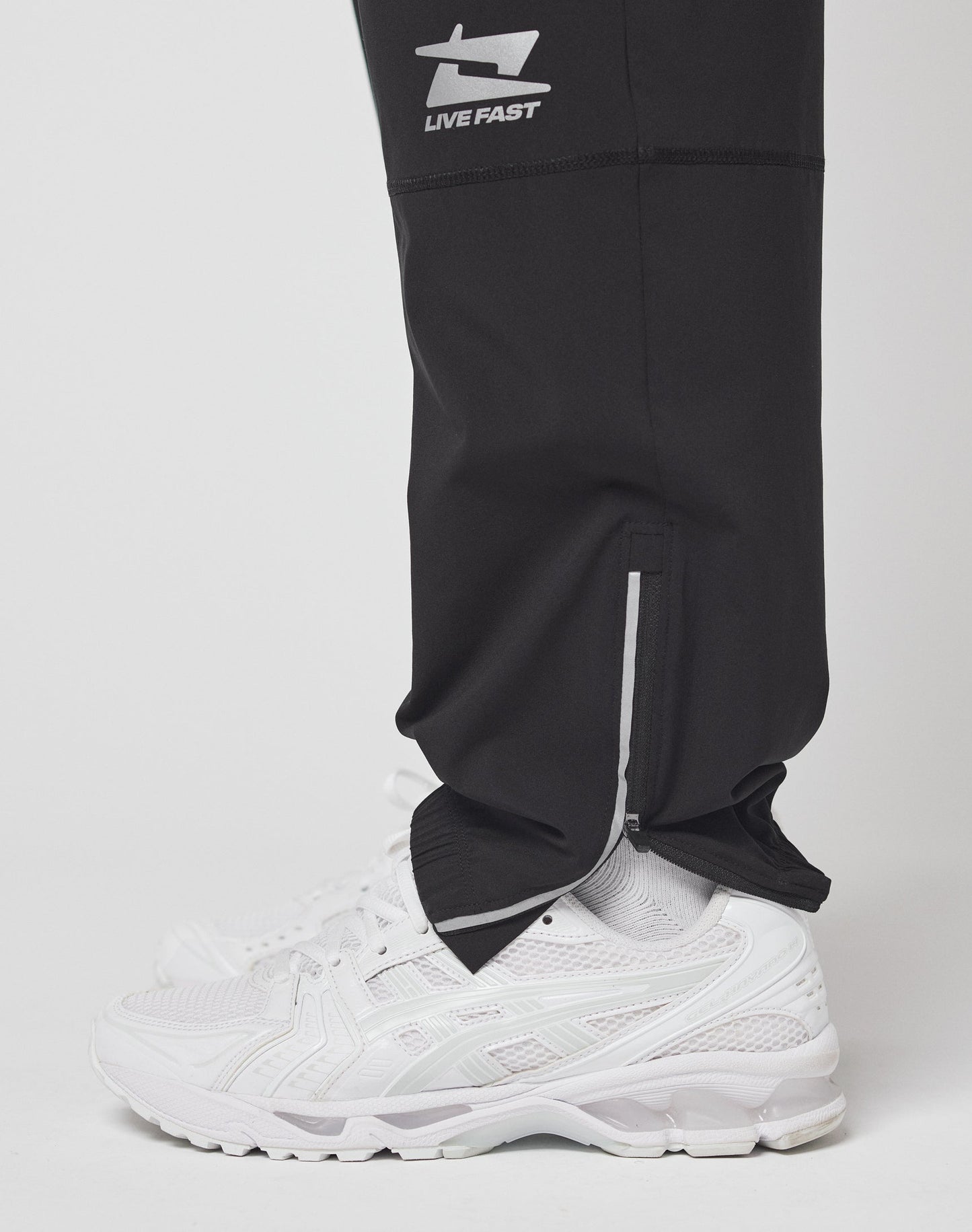 Side view of a person in LIVE FAST DIE YOUNG Live Fast Performance Running Pants, featuring an elastic waistband, ankle zipper, LIVE FAST logo, and reflective print near the knee. Paired with white sneakers. Background is plain white.