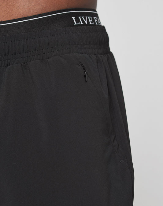 Close-up of a person wearing LIVE FAST DIE YOUNG Live Fast Performance Running Pants with an elastic waistband, small side zip pocket, and reflective print for added style. The waistband shows the LIVE F... logo partially visible.